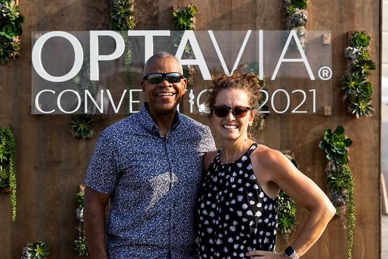 A male and female attendee posing together in front of a wooden backdrop with Optavia logo and succulent plants, smiling for the camera.